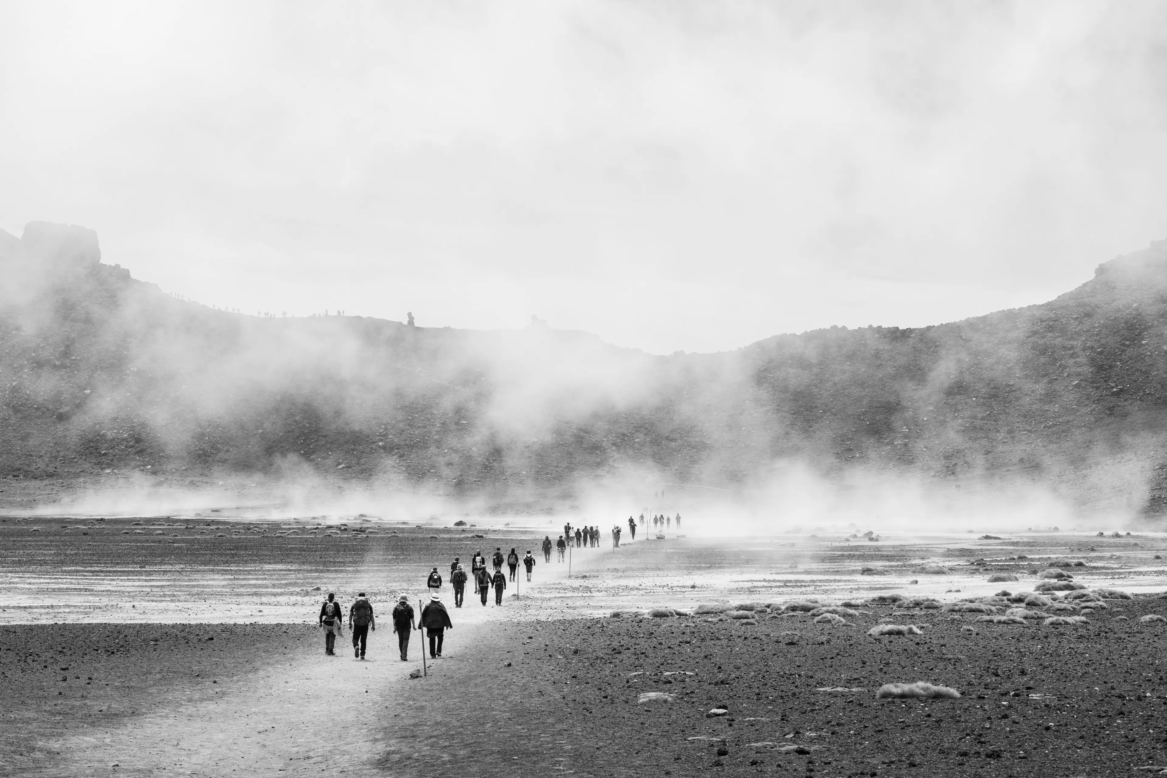 A black and white photo of several people walking along a barren landscape towards a mountain covered in mist.
